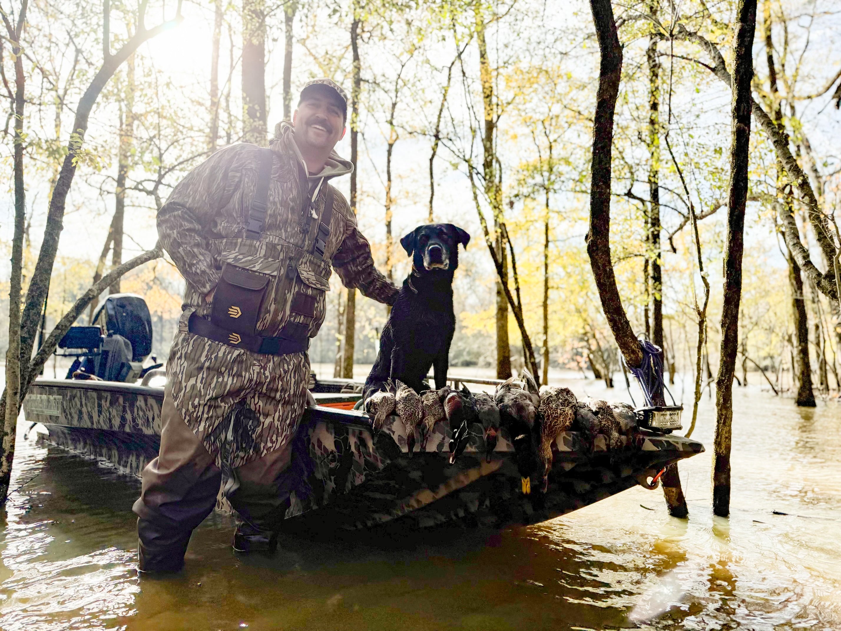 Hunter standing on a boat in flooded timber with a black lab and harvested ducks at sunrise.